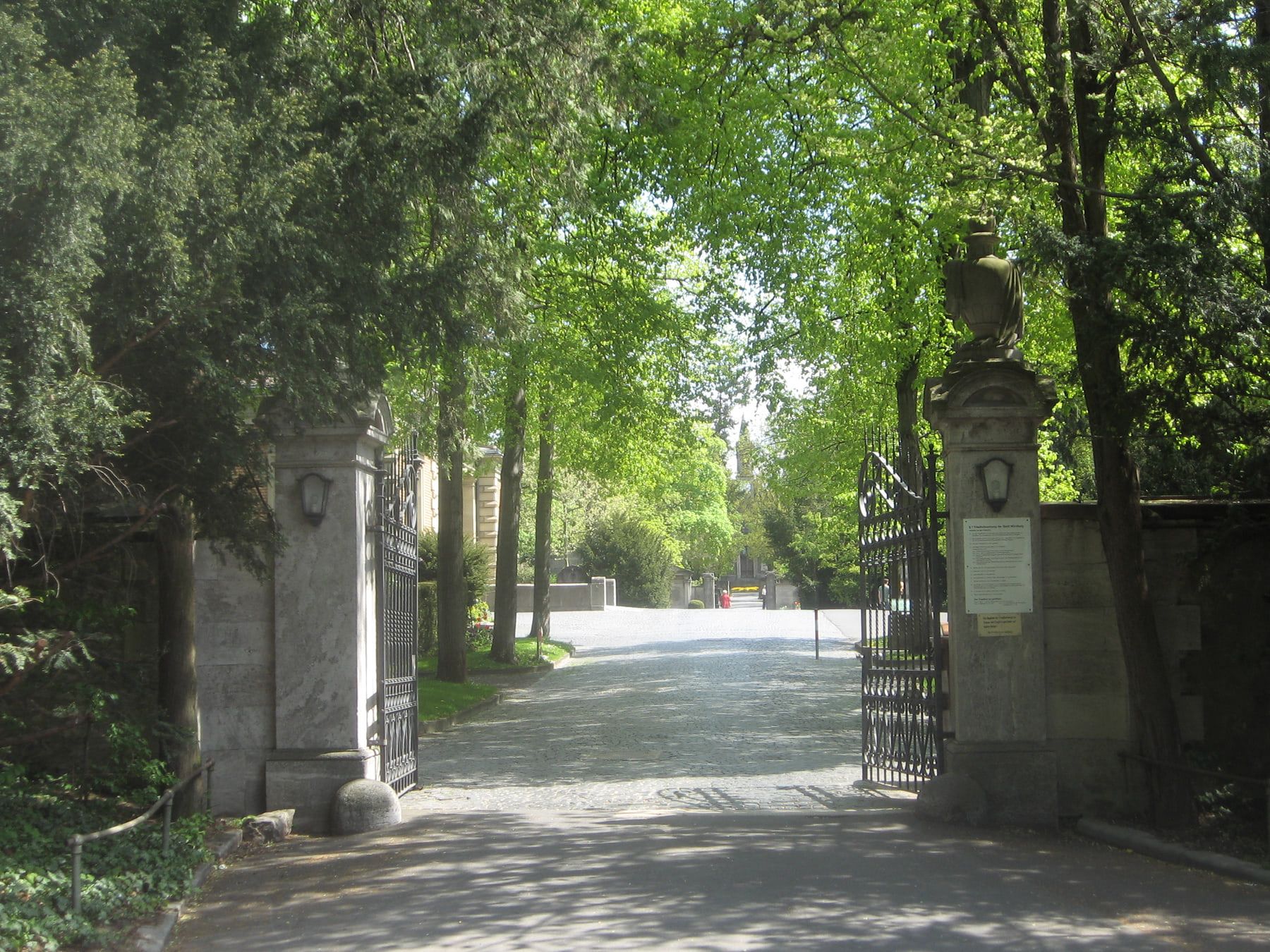 Geöffnetes schmiedeeisernes Haupttor zwischen Steinpfeilern mit Blick in die baumbestandene Hauptallee auf Hauptfriedhof Würzburg
